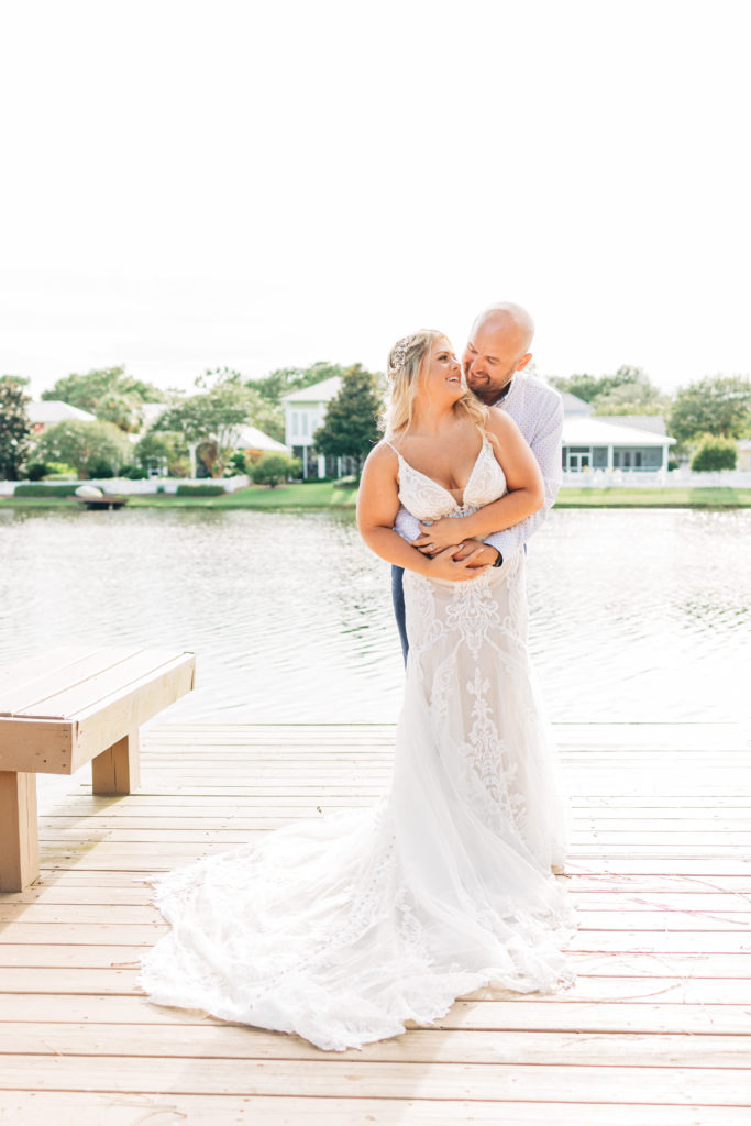 Groom hugs bride from behind while they smile on a dock by the water at Sandestin Resort in Miramar Beach Florida